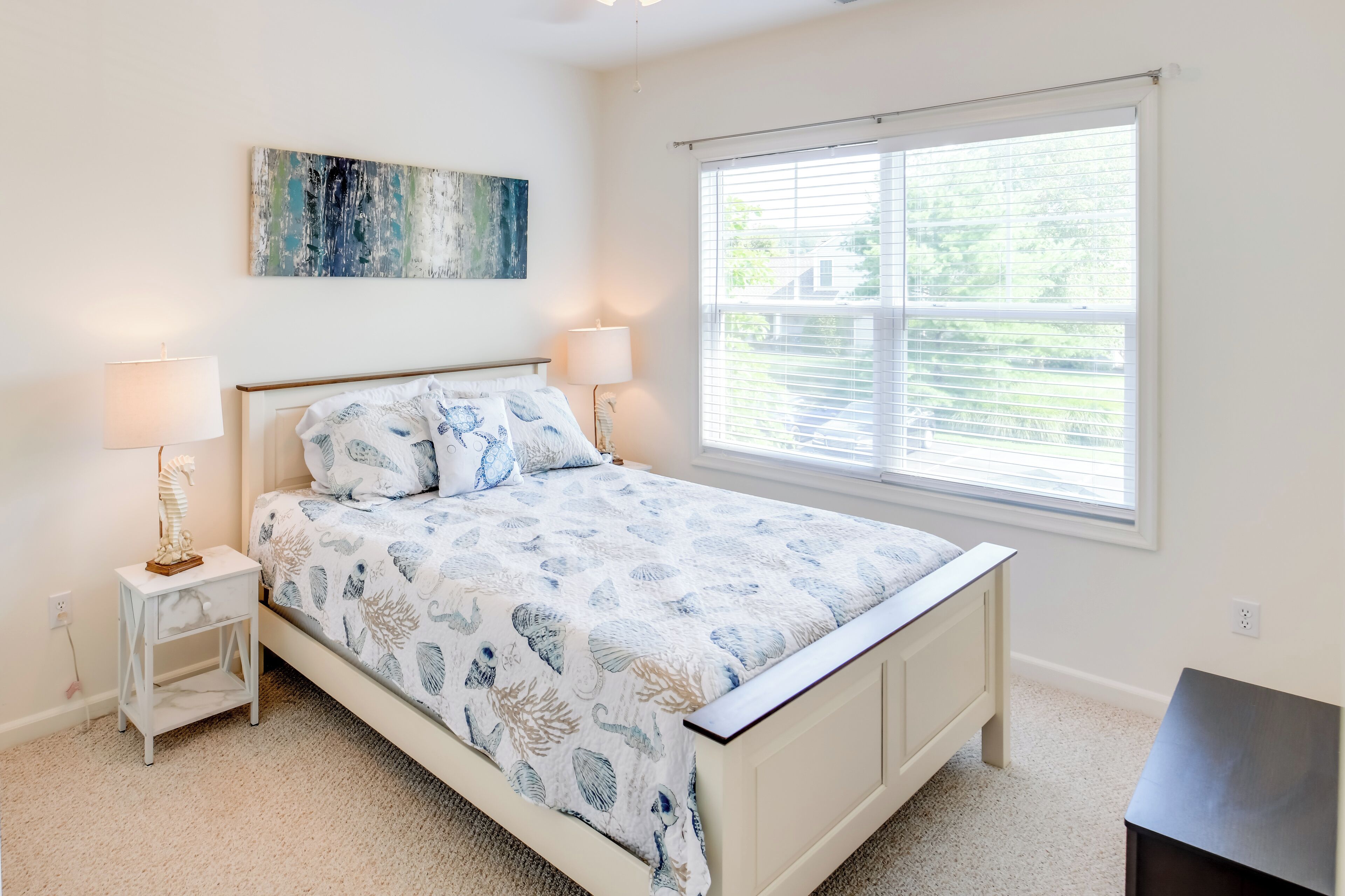 A bedroom with a bed, two bedside lamps, a nightstand, a window with blinds, and a painting on the wall.