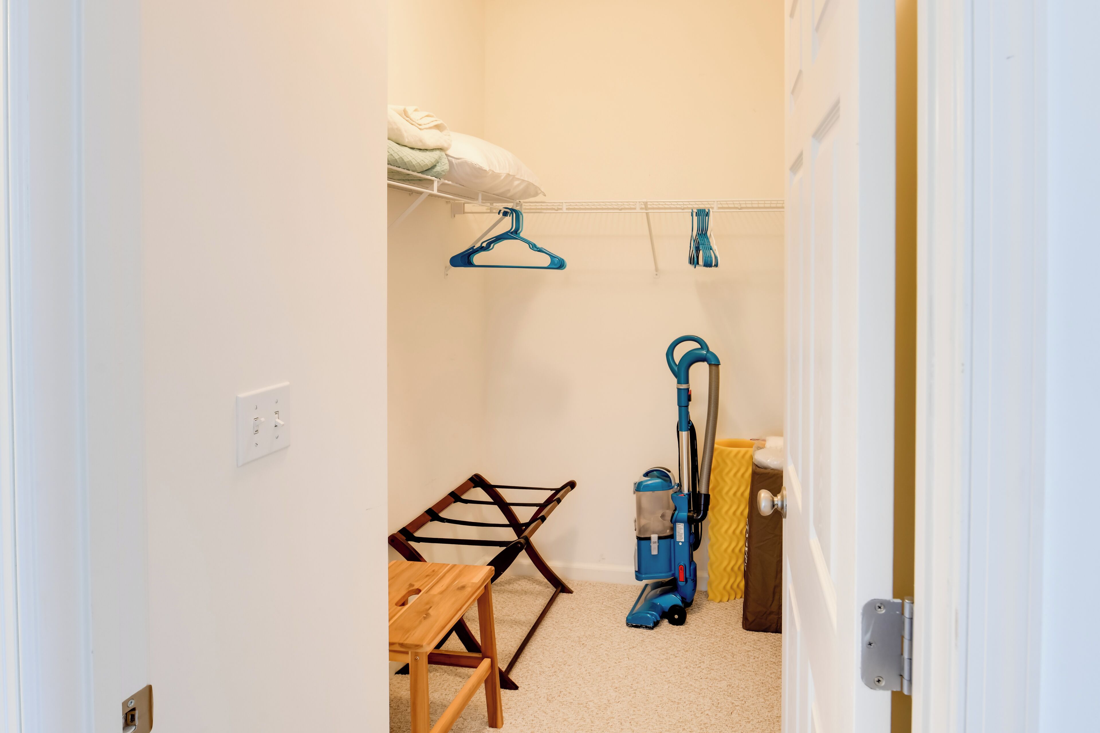 Una habitación con una mesa de madera, un perchero, un aspirador y una cama con un almohábil blanco.