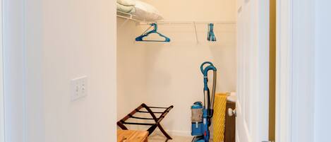 Una habitación con una mesa de madera, un perchero, un aspirador y una cama con un almohábil blanco.