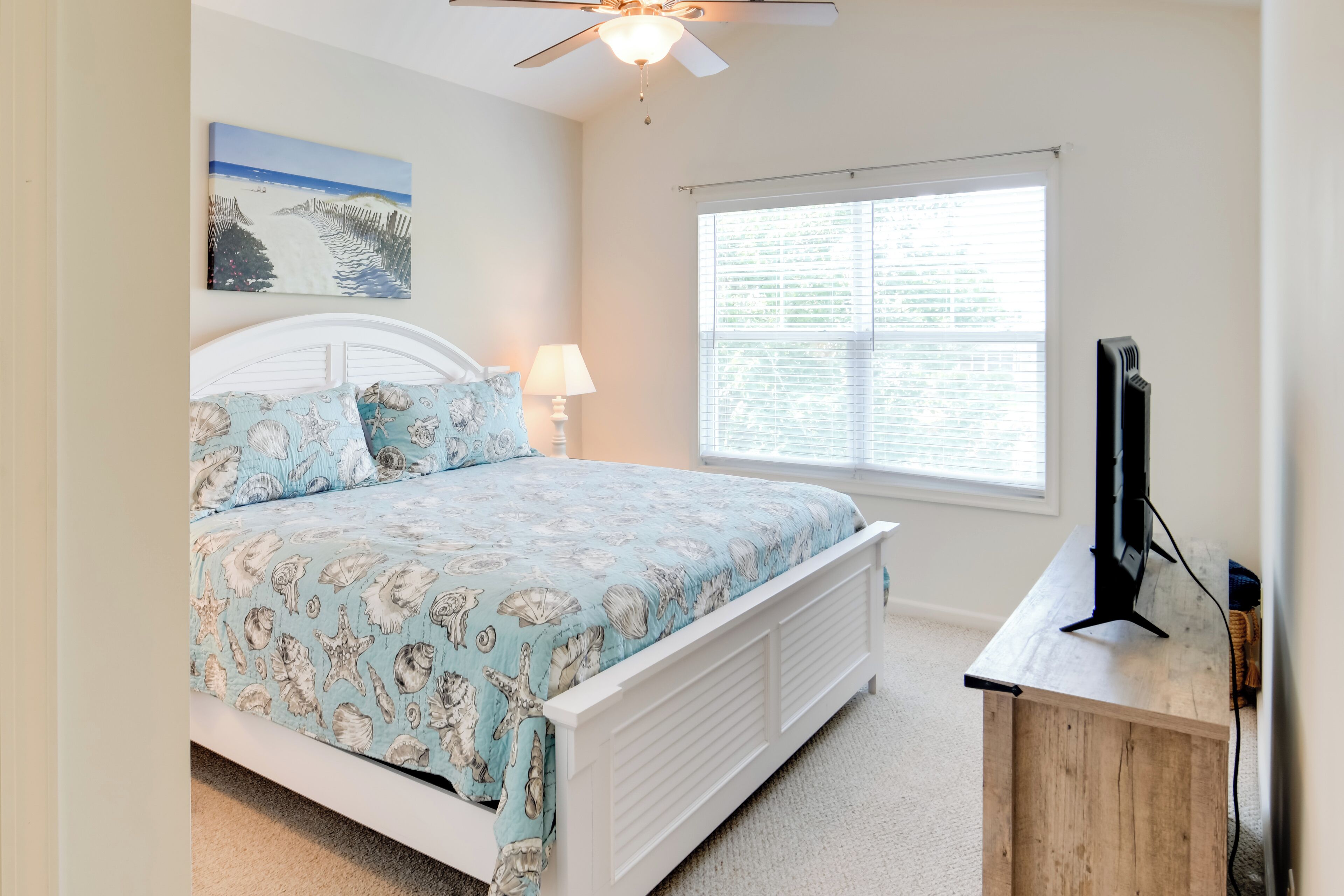 A bedroom with a bed, a ceiling fan, a window with blinds, and a TV on a wooden stand.