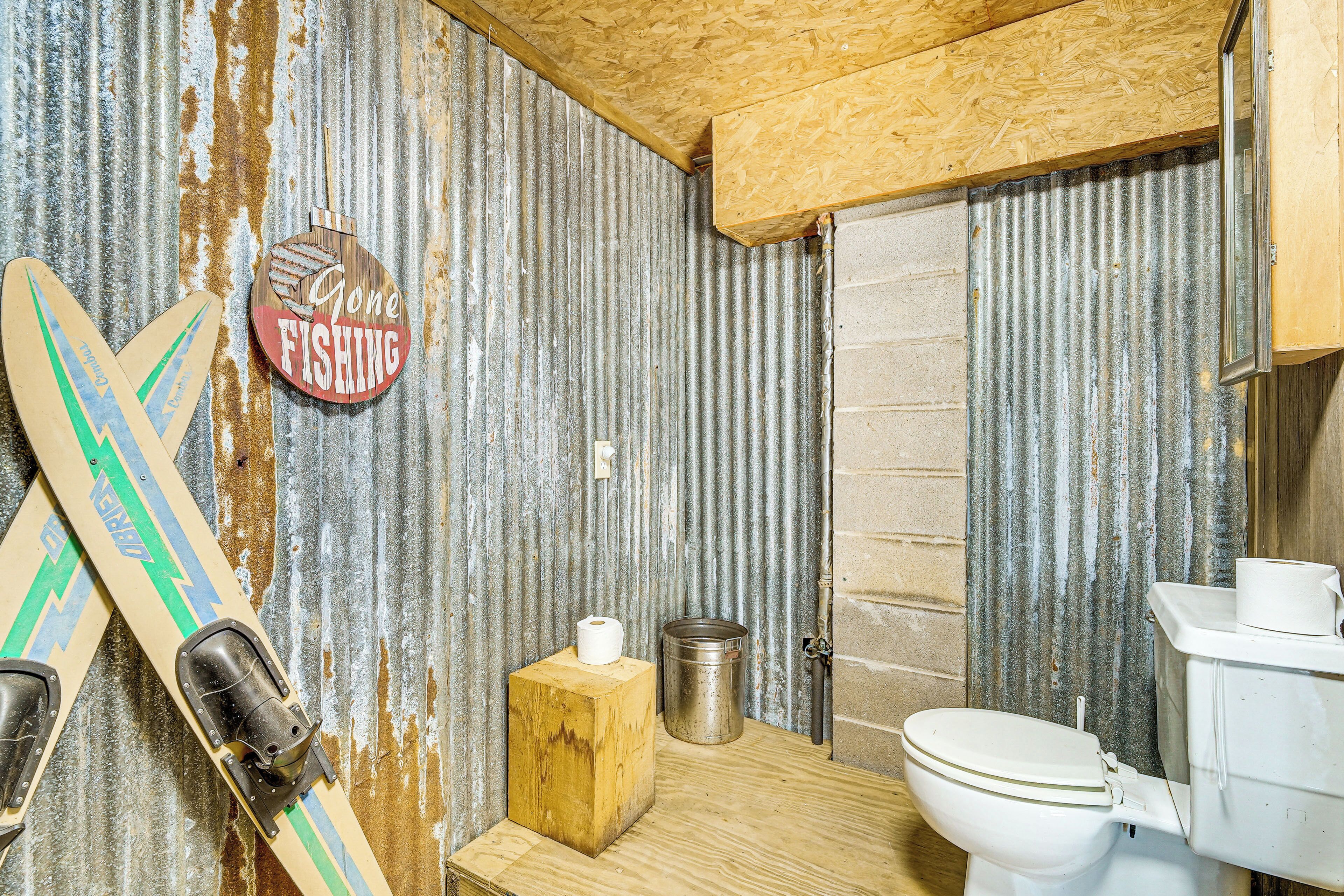 A rustic bathroom with a wooden floor, a toilet, a metal bucket, and a sign that says "Gone Fishing".
