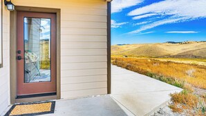 Uma casa com porta marrom, varanda de concreto e vista para um campo gramado e colinas.