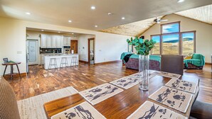 Uma sala de estar ampla com cozinha, mesa de jantar com jogos americanos e uma grande janela com vista para a paisagem.