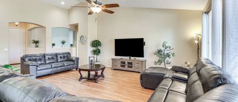 A spacious living room with leather sofas, a flat-screen TV, wooden flooring, and a ceiling fan.