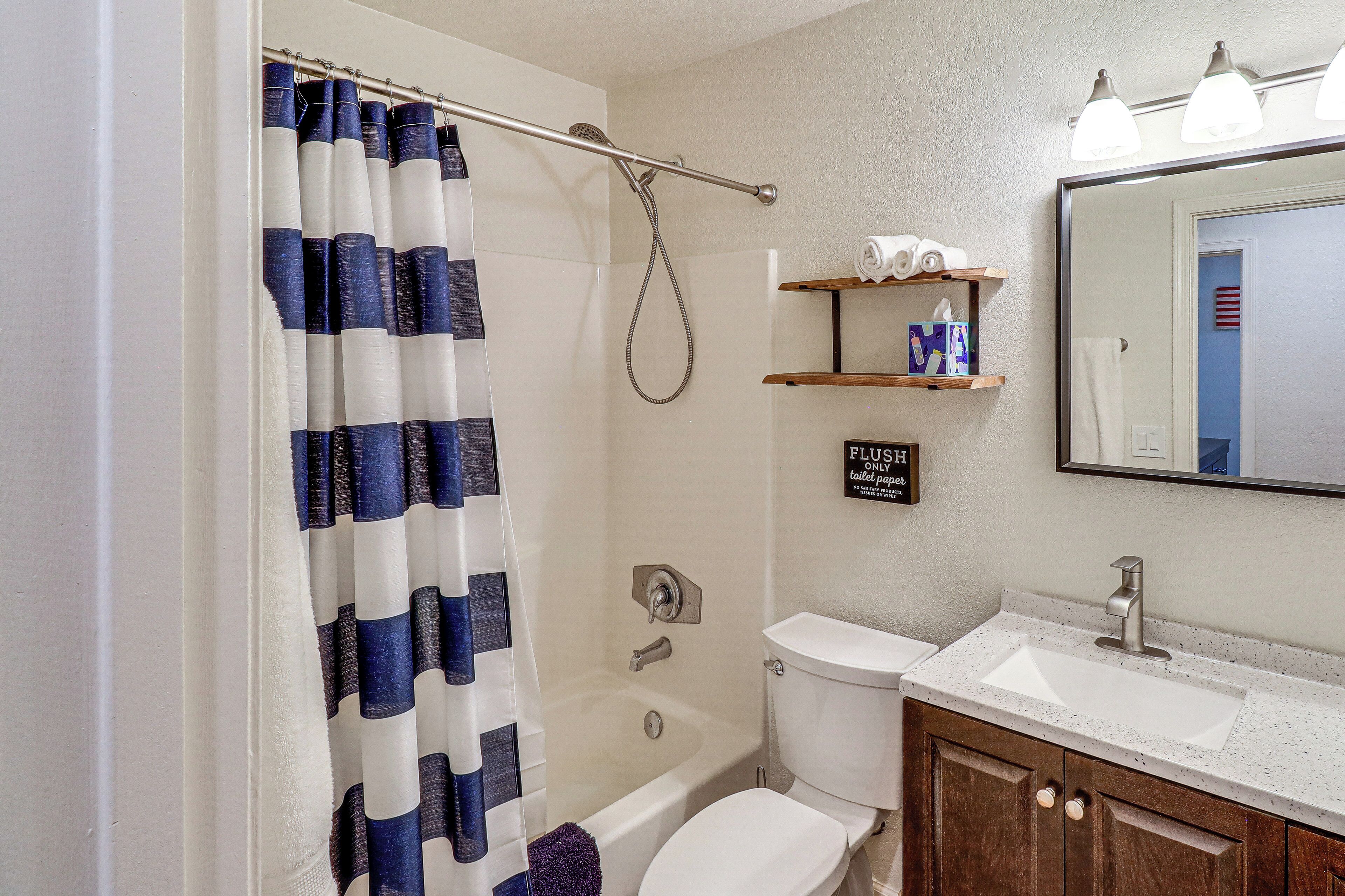 A bathroom with a shower curtain, bathtub, toilet, sink, and a shelf with toiletries.