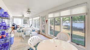 Un patio soleado con sillas y una mesa blancos, ventanas amplias con persianas y un ventilador de techo.