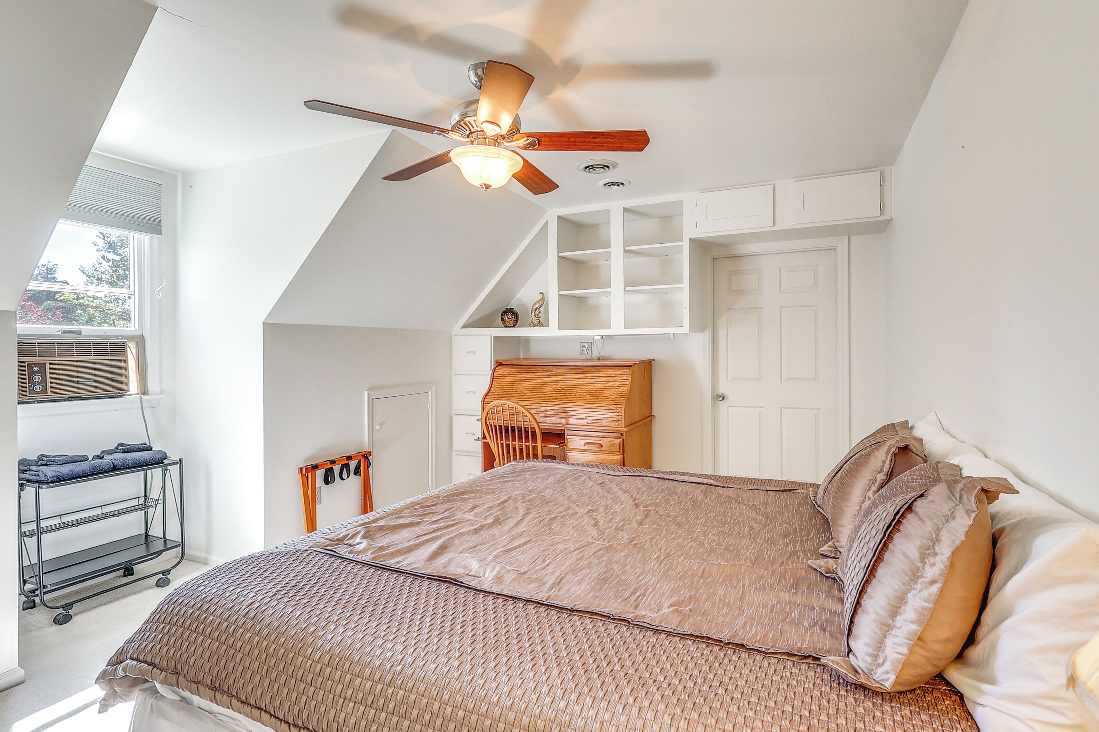 A bedroom with a sloped ceiling, a ceiling fan, a bed with a brown bedspread, a wooden desk, and a window with blinds.