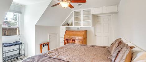 A bedroom with a sloped ceiling, a ceiling fan, a bed with a brown bedspread, a wooden desk, and a window with blinds.