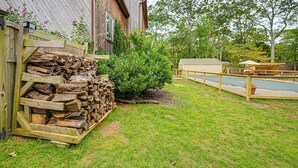 Un bâtiment en bois, avec une pile de bois de chauffage, un petit buisson et une piscine entourée d’une clôture.