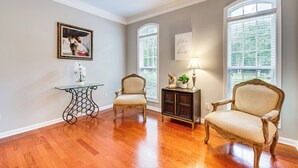 A room with hardwood flooring, two upholstered chairs, a glass table with a vase of flowers, a dark wooden cabinet, and a framed picture on the wall. - 3 Mi to Avalon: Townhome w/ Balcony in Alpharetta! (Milton)