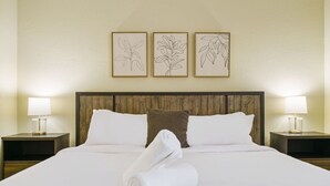 Un letto rifatto con lenzuola bianche, una testiera in legno e un cuscino decorativo marrone. Due comodini con lampade e tre stampe botaniche incorniciate sulla parete sopra il letto.