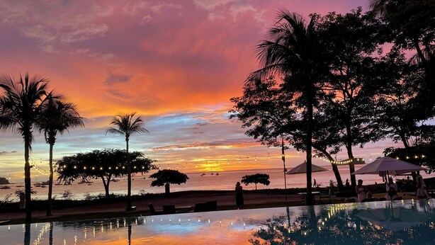 Une vue sur la piscine, avec un coucher de soleil, des palmiers et une mer calme.
