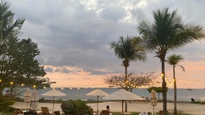 Un espace aménagé au bord de la piscine, avec des chaises longues et des parasols, une petite piscine et une vue sur la mer, où l’on peut apercevoir des bateaux.