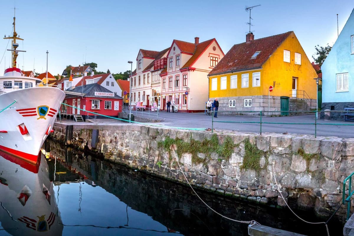 En havn med både, en stenvæg og farverige bygninger, heriblandt en bygning i rødt og hvidt med teksten "CHRISTIANSO".