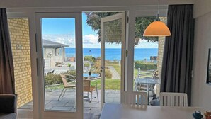 Habitación con puerta de vidrio corredera que da a un patio exterior con vista al mar. Hay una mesa y sillas blancas, y una lámpara colgante.