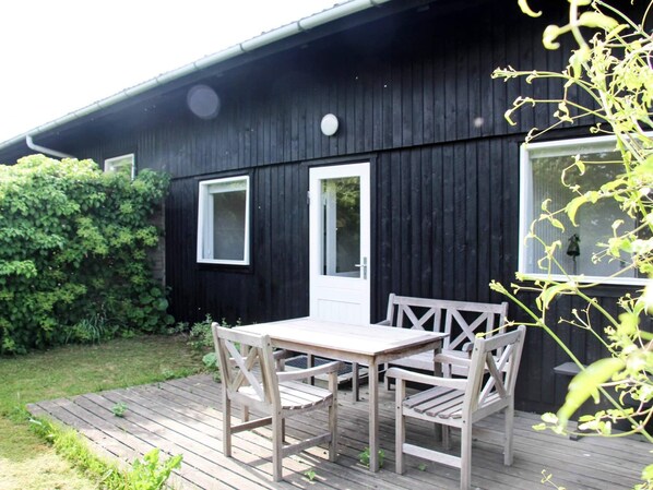 A wooden deck with a table and chairs, a black building with two windows, and a white door. - 4 Person Holiday Home in Svaneke (Svaneke)