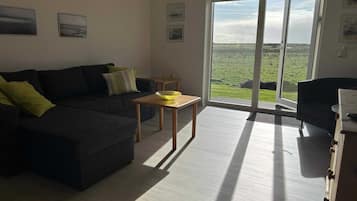 Una sala de estar con un sofá de color oscuro, una mesita de madera con un cuenco amarillo, y vista a un paisaje verde a través de grandes puertas de vidrio.