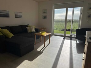 A living room with a dark-colored sofa, a small wooden table with a yellow bowl, and a view of a green landscape through large glass doors.
