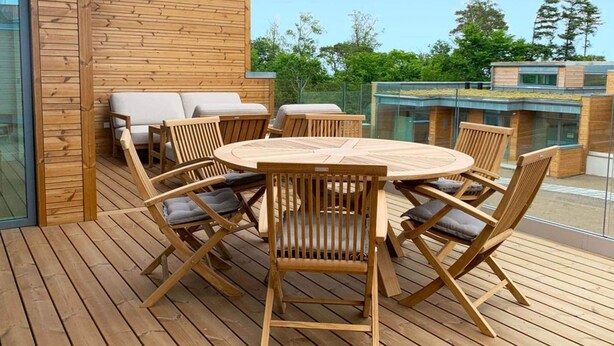 A wooden deck with a round table and chairs, a sofa, and a glass railing. - 6 Person Holiday Home in Svaneke (Svaneke)