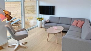 A modern living room with a grey sofa, a small round table, a single chair, a TV mounted on the wall, and a large window with a view of a wooden deck. - 6 Person Holiday Home in Svaneke (Svaneke)