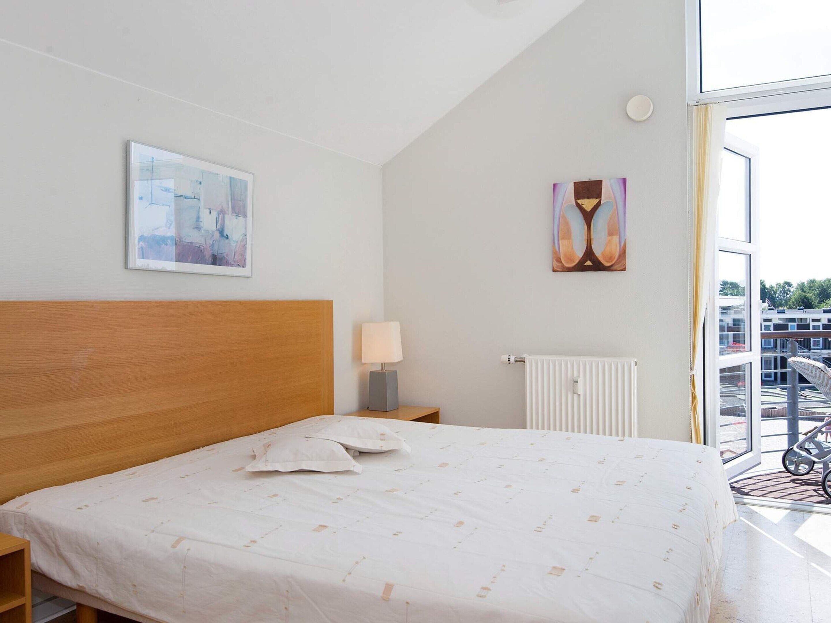 Habitación de hotel con cabecera de madera, cama con cubrecama blanco, mesita de noche con lámpara, un cuadro en la pared y ventana con vistas a edificios.