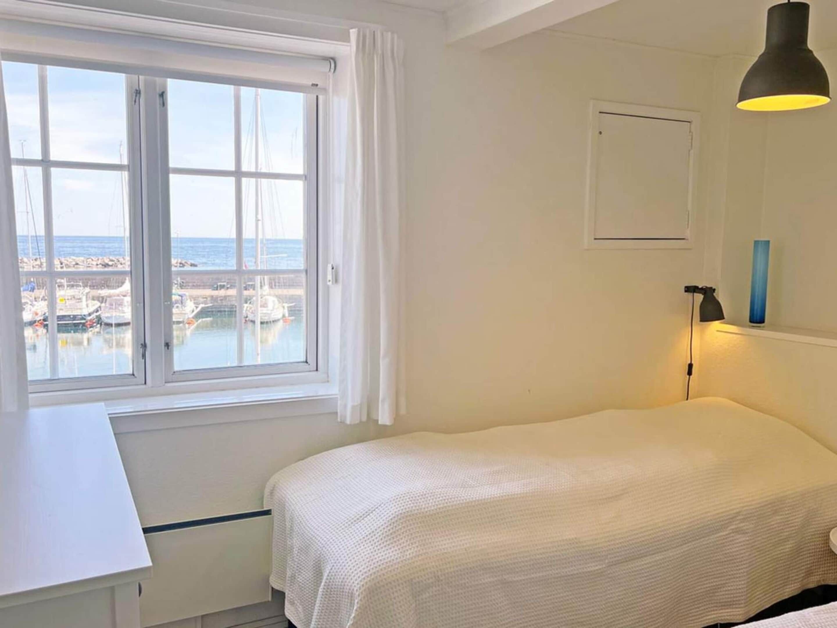 A bedroom with a bed, a desk, a window with a view of boats and water, a lamp, and a framed picture on the wall.