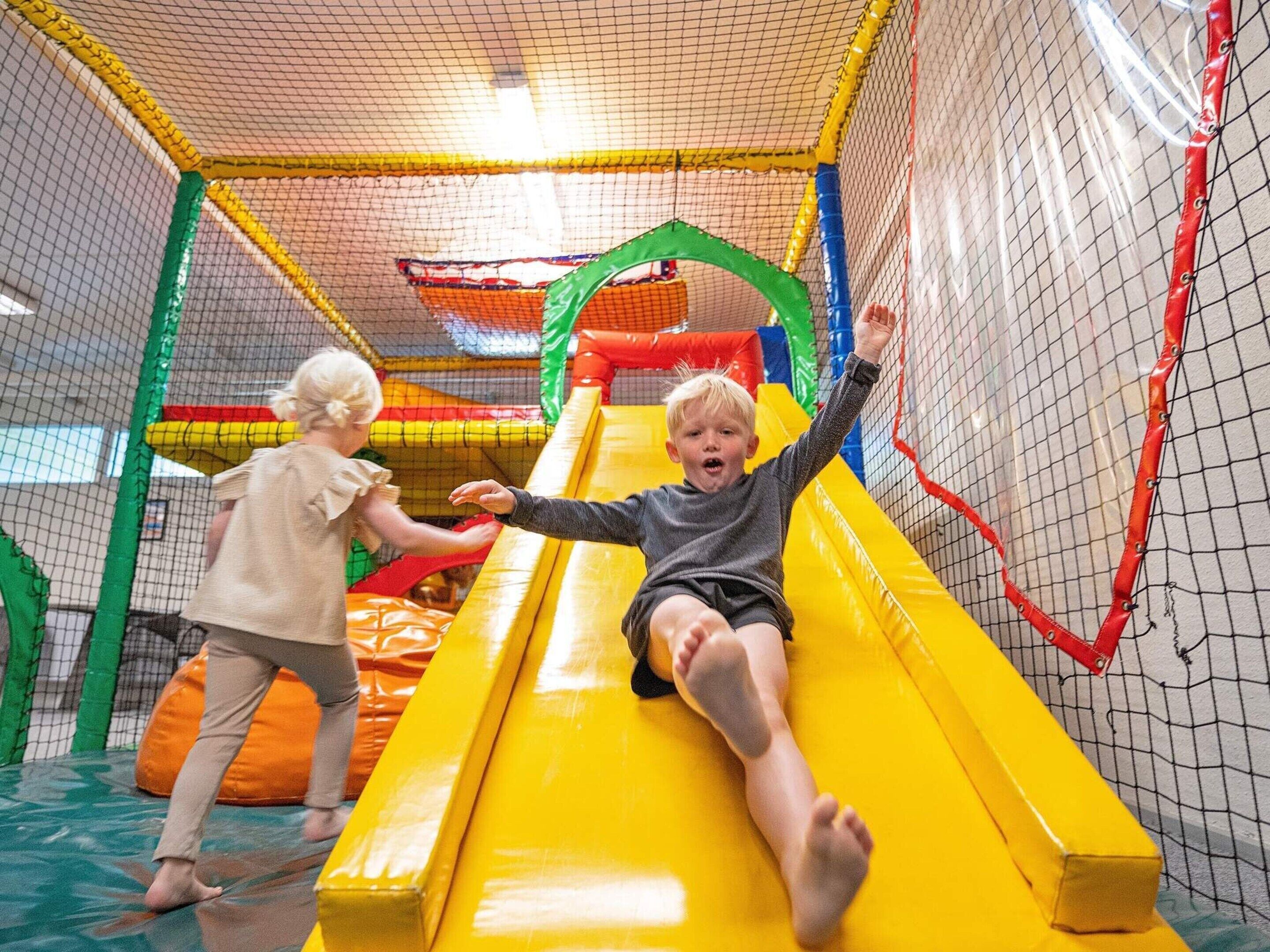 Children playing on a yellow slide inside a play area with netted walls and colorful play structures.