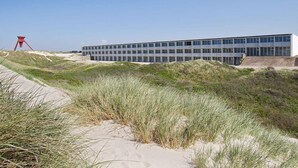Ein modernes Gebäude mit flachem Dach und großen Fenstern, umgeben von Sanddünen und Gras.