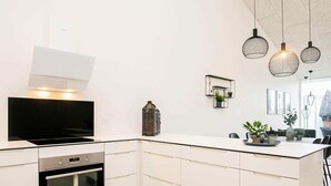 Une cuisine moderne avec des meubles blancs, un four encastré et un grand îlot central doté d’un évier. Des luminaires suspendus sont placés au-dessus de l’îlot, et une petite lanterne décorative est posée sur le plan de travail.