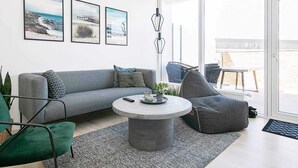 Un salon moderne avec un canapé gris, une table basse ronde et un fauteuil gris. Trois tableaux encadrés ornent le mur, ainsi qu’un luminaire suspendu. De grandes fenêtres offrent une vue sur l’extérieur.