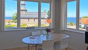 Zona pranzo con tavolo e sedie, ampie finestre vista su un edificio e il mare.