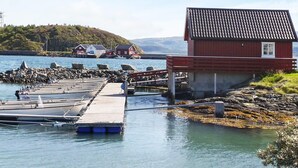 Escena costera con un muelle de madera que se adentra en aguas tranquilas, un edificio rojo con techo de tejas y un fondo de colinas y casas.
