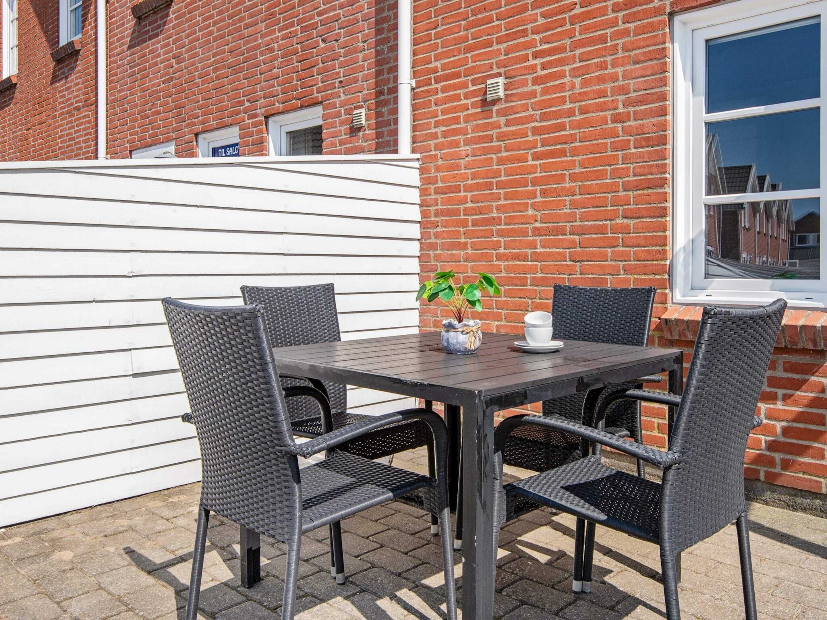 An outdoor patio with a table and four chairs, a potted plant, and a window with a view of a street.