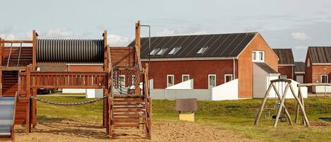 Une zone de plage sablonneuse avec une structure de jeux en bois, une balançoire et un bâtiment en briques doté d’un toit en pente.