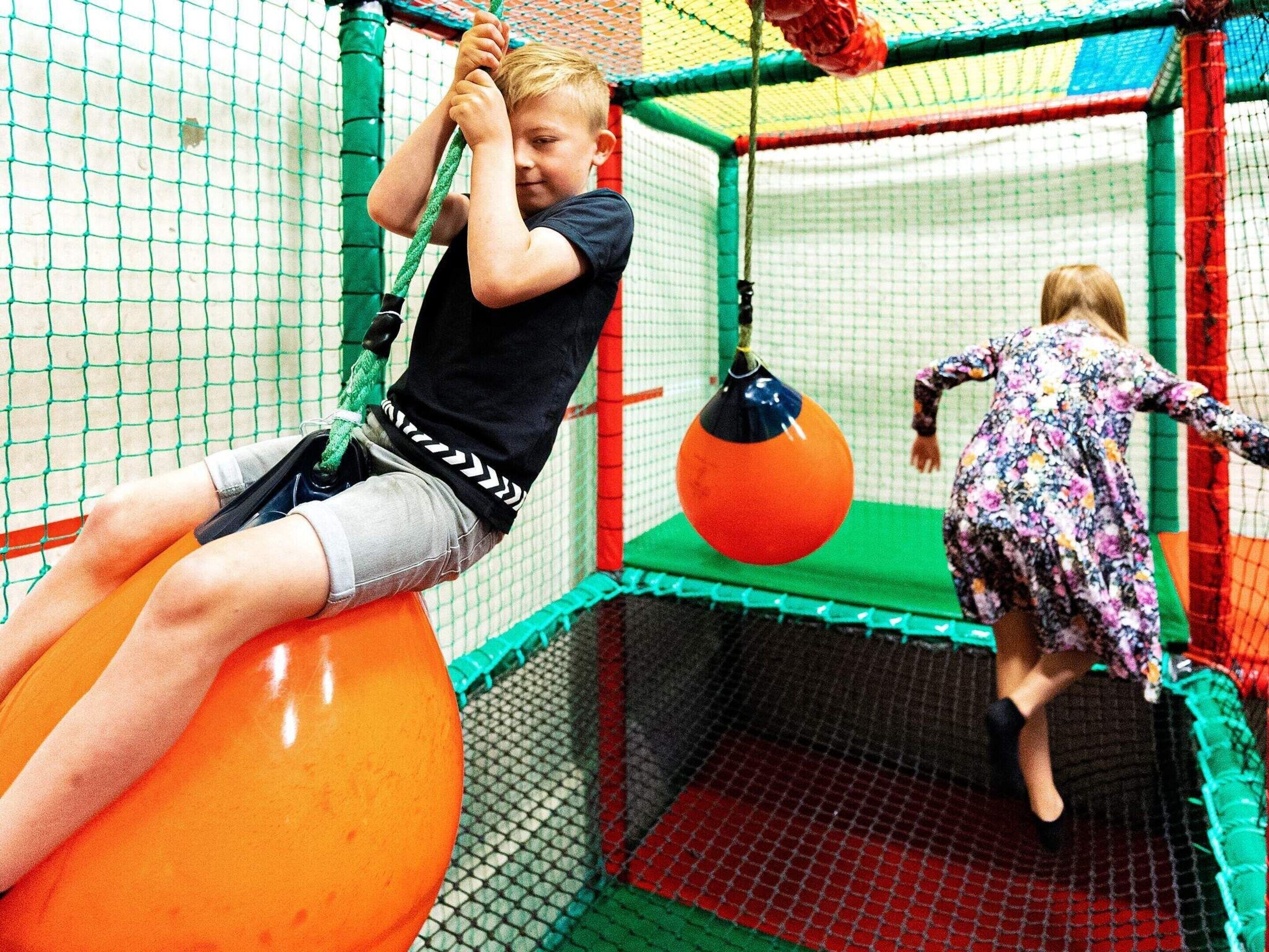 A child climbing on a net structure with a ball and another child in a floral dress.