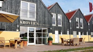 Un edificio con el letrero "KROYER'S GL. SKAGEN", que presenta una entrada amplia, asientos exteriores con mobiliario de mimbre y una bandera roja en un asta.