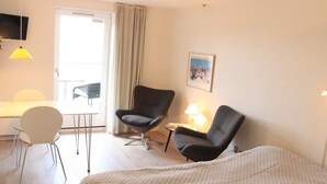 Hotellihuone, jossa on sänky, kaksi nojatuolia, työpöytä, tuoli, televisio ja taulu seinällä.
