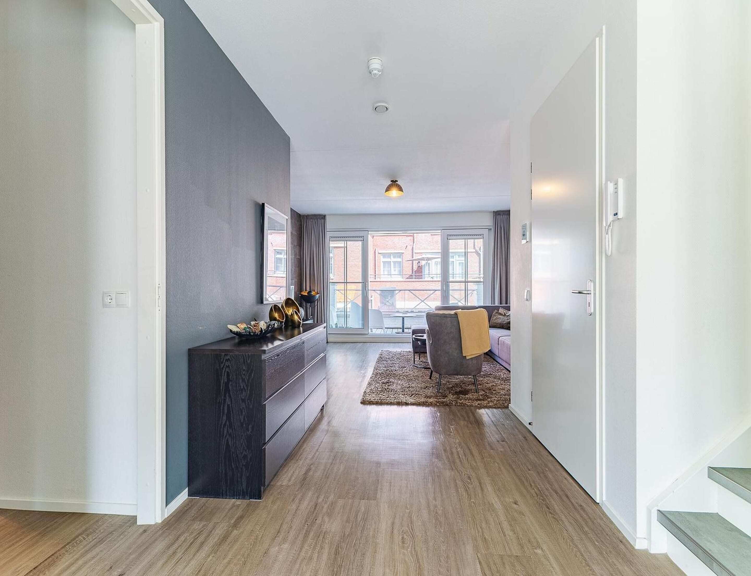 A modern living room with a dark wood floor, a grey accent wall, and a large window with a view of a brick building.