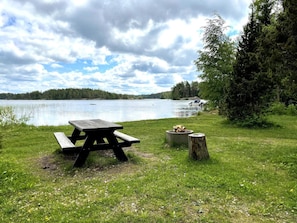 Ein Picknicktisch mit Feuerstelle, eine Rasenfläche und ein See mit Booten im Hintergrund.
