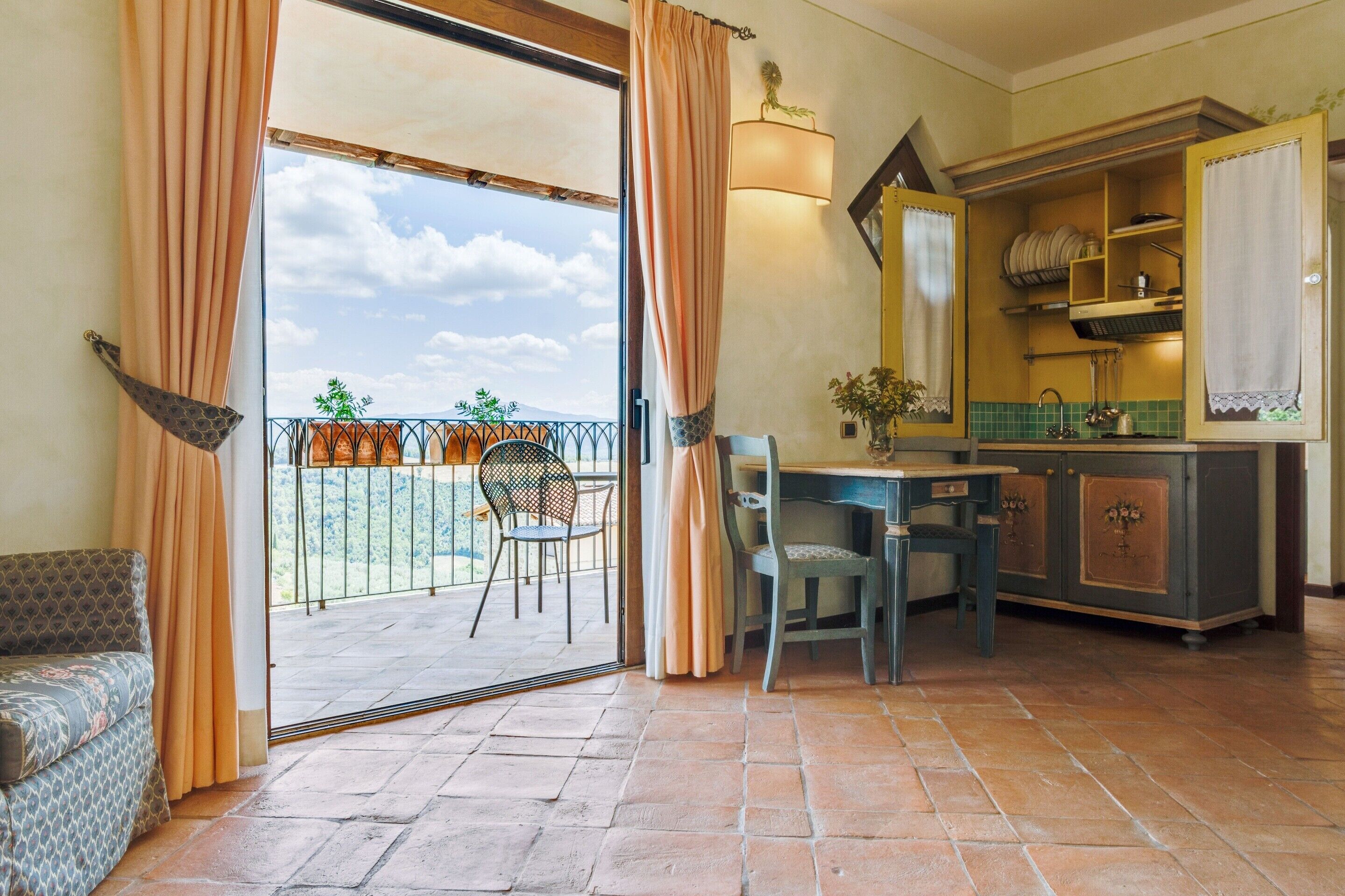A room with a balcony, a dining table with chairs, a sofa, and a kitchen area with cabinets and a sink.