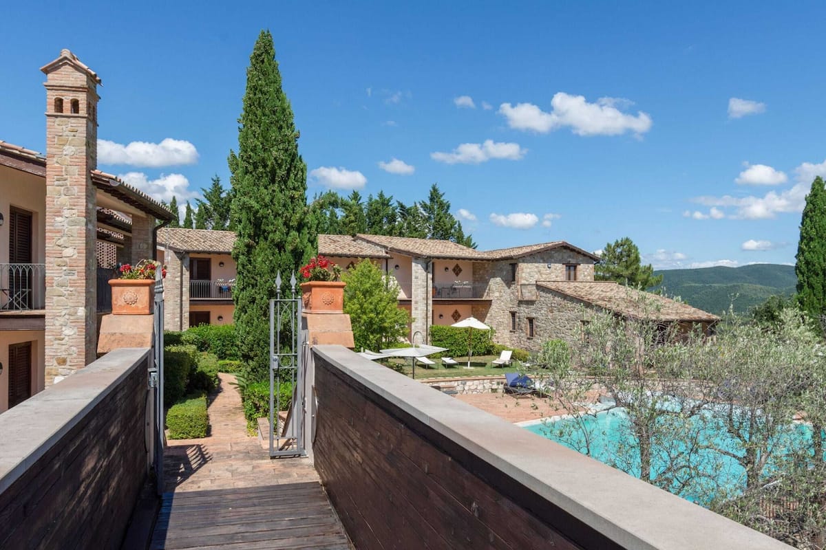 A resort with a wooden deck leading to a pool area, stone buildings, and a clear view of the mountains.