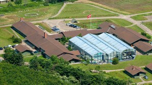 Vue aérienne d’un complexe immobilier moderne composé de plusieurs bâtiments, dont une grande structure entièrement vitrée abritant une piscine, le tout entouré de places de stationnement et d’espaces verts.