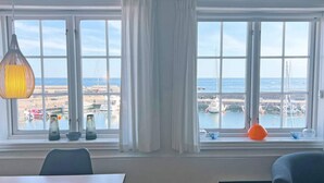 Une pièce dotée d’une grande fenêtre donnant sur un port de plaisance et une plage, et comprenant une table, des chaises et des objets décoratifs.