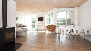 A modern living room with a fireplace, wooden ceiling, large windows, and a dining area with white furniture. - 10 Person Holiday Home on a Holiday Park in Blavand (Blavand)