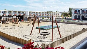 En legeplads med gyngestole og trampolin, omgivet af et sandareal og en bygning med flere altaner.