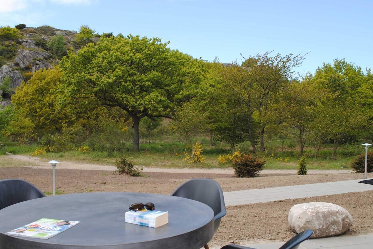 An outdoor seating area with a round table and chairs, surrounded by trees and a dirt path.