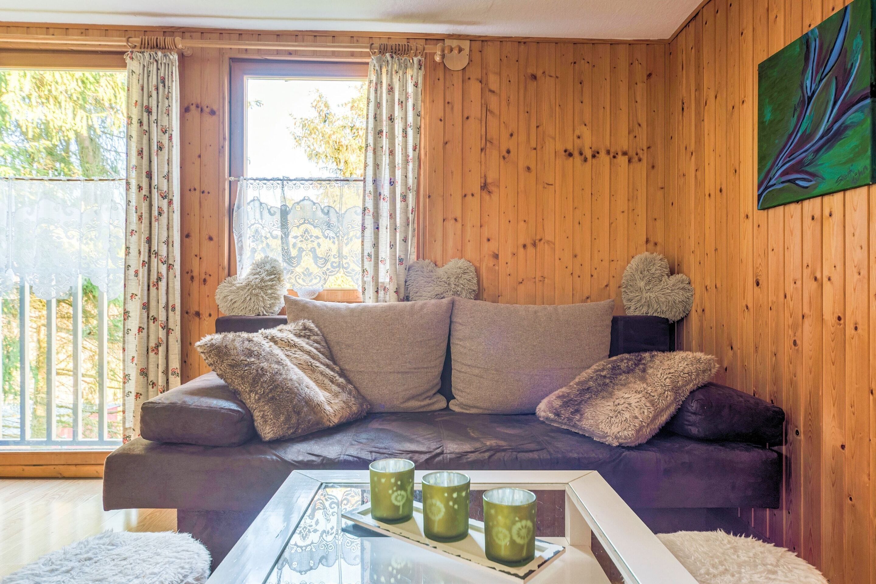 A cozy room with a wooden paneled wall, a cushioned sofa with fluffy pillows, and a glass coffee table with three green candles.