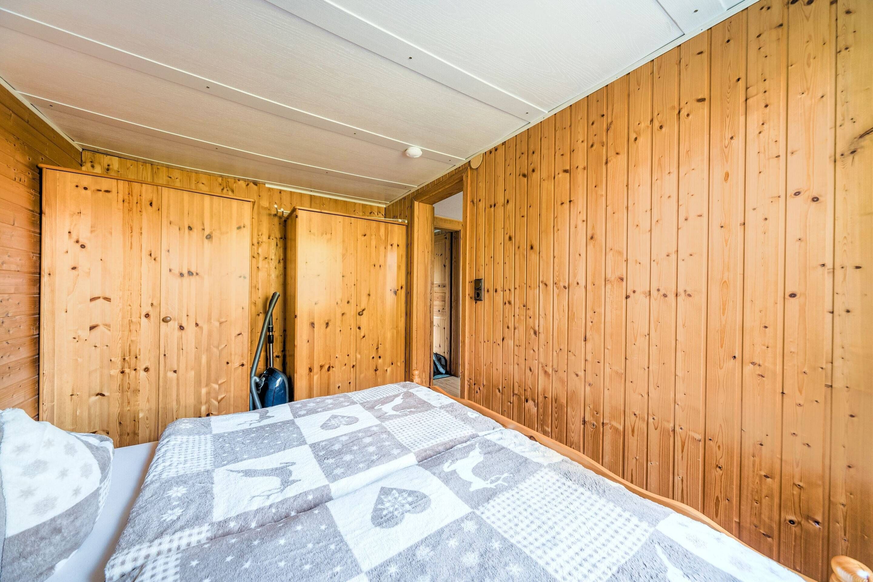A wooden-paneled room with a bed, a vacuum cleaner, and a door leading to another room.