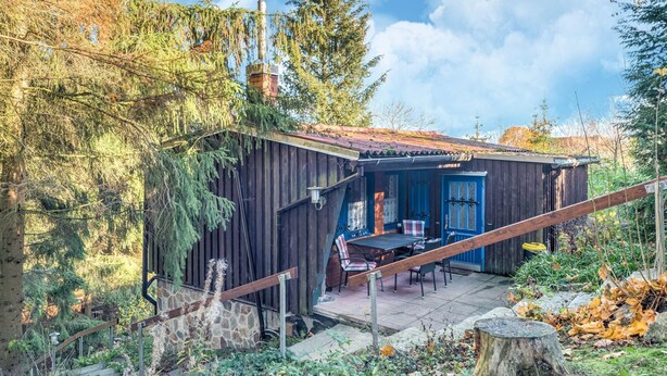 A rustic cabin with a wooden exterior, a chimney, a dining area with outdoor seating, and a stone staircase leading to the entrance. - Comely Holiday Home in Guntersberge Near Forest (Harzgerode)