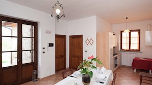 A dining area with a table set for two, a hanging light fixture, wooden doors, and a window with curtains. - Stunning Farmhouse in Passignano With Pool (Passignano sul Trasimeno)